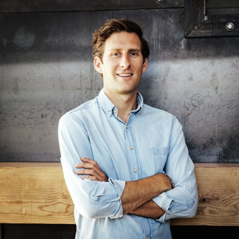 The image shows a man with brown hair and blue eyes, wearing a light blue button-down shirt. He is standing with his arms crossed, leaning against a wooden surface. The background appears to be a textured gray wall. The man has a friendly expression and seems to be posing for the photo.